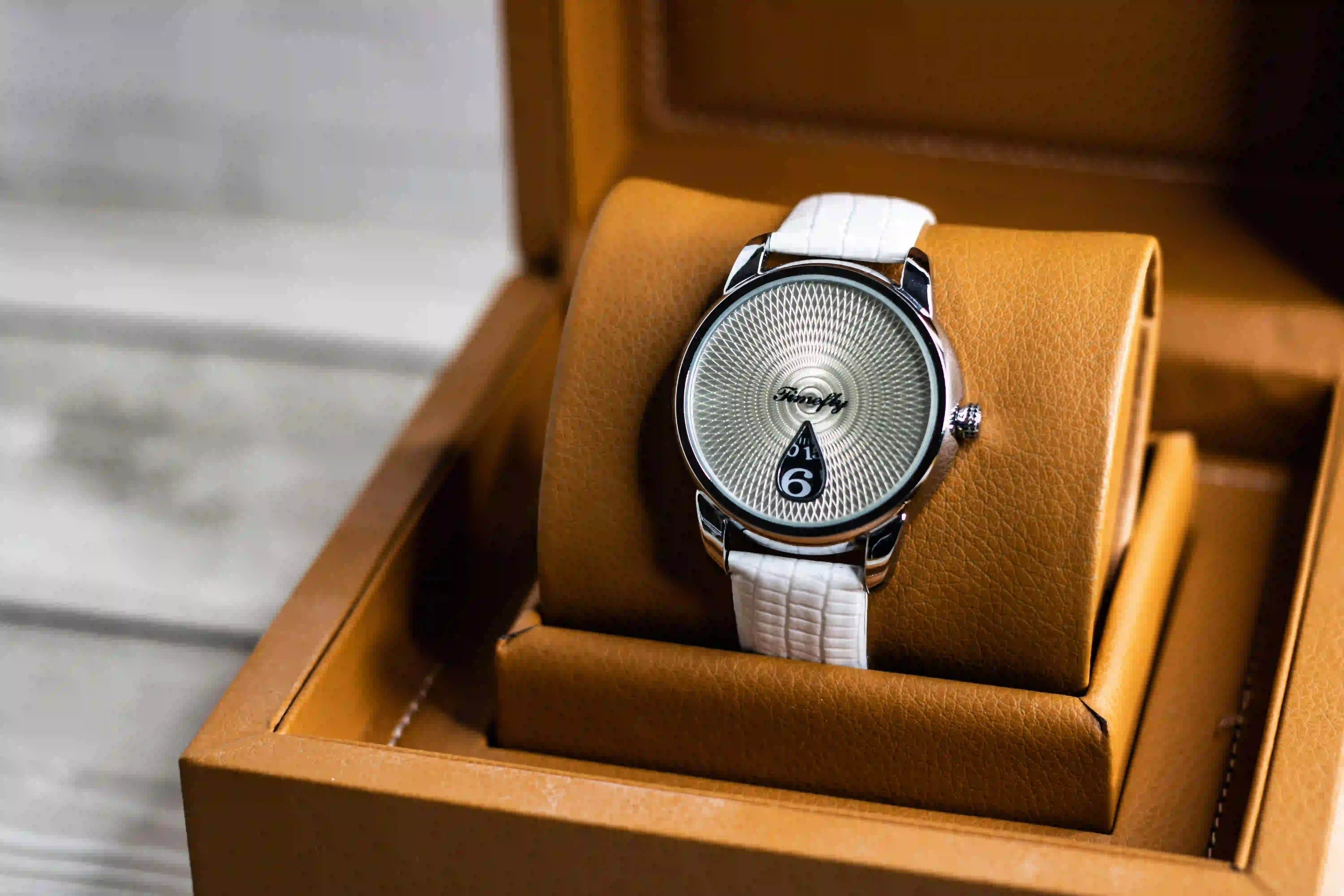 Reloj pequeño unisex con correa blanca y esfera plateada en caja de cuero marrón.
